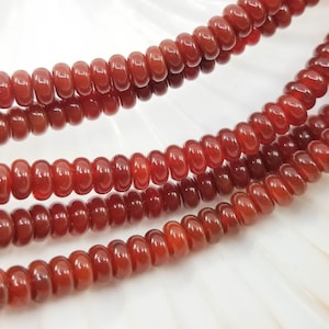 Peut inclure: Gros plan sur des brins de perles de pierres précieuses polies, de couleur brun rougeâtre. Les perles sont de taille uniforme, de forme arrondie et en forme de disque. Les perles ont une finition brillante et sont enfilées ensemble.