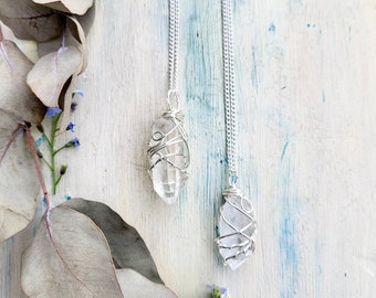 May include: Two clear quartz crystal pendants wrapped in silver-colored wire, suspended from delicate silver chains. The crystals are displayed against a backdrop of a white and blue painted surface, with eucalyptus leaves and small blue flowers.