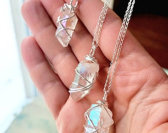 Puede incluir: Tres collares de cristal de cuarzo transparente con destellos iridiscentes de color. Cada cristal está envuelto en alambre plateado y cuelga de una delicada cadena plateada. Los collares se muestran sobre un fondo suave y borroso.