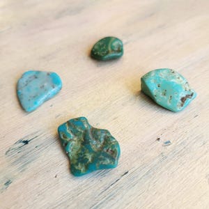 Può includere: Quattro pietre di turchese naturali in varie tonalità di blu e verde sono disposte su una superficie di legno chiaro. Le pietre hanno forme e texture irregolari, che ne evidenziano la bellezza naturale.