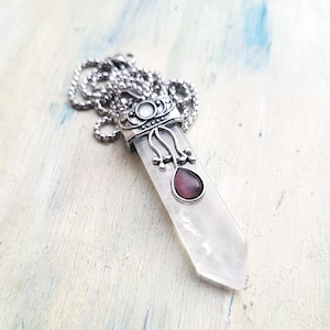 May include: A silver-toned pendant necklace featuring a clear, elongated crystal with a purple gemstone accent. The pendant is suspended from a silver chain. The design includes ornate metalwork around the crystal and gemstone.