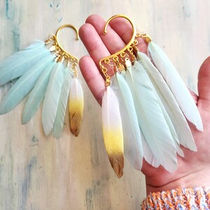 Può includere: Polisini per orecchie color oro adorni di piume azzurre e bianche. Le piume hanno una sfumatura dorata alle punte. I polsini per orecchie sono progettati per avvolgere l'orecchio.