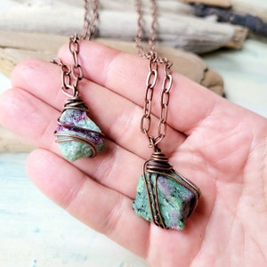 Puede incluir: Dos colgantes de piedra preciosa verde y rosa envueltos en alambre de cobre en cadenas de cobre. Los colgantes cuelgan de la mano de una persona.