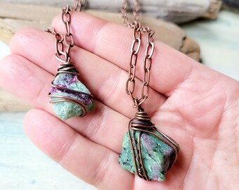 May include: Two green and pink gemstone pendants wrapped in copper wire on copper chains. The pendants are hanging from the hand of a person.
