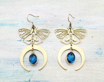 Puede incluir: Pendientes de tono dorado con un diseño de media luna y una piedra preciosa azul colgante. La media luna está coronada con un diseño de mariposa recortada.