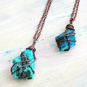 May include: Two turquoise gemstone pendants wrapped in copper wire, hanging from copper chains. The pendants are different sizes and shapes.