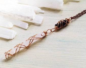 May include: A necklace featuring a long, pale pink crystal point and a smaller, dark black crystal, both wrapped in copper wire. The crystals are connected by a copper link and attached to a dark brown metal chain, creating a layered design.