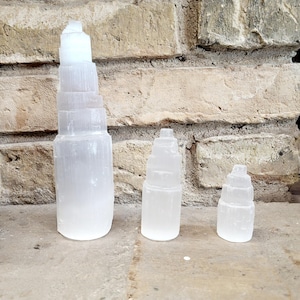 Op de afbeelding: Drie witte seleniet torensculpturen van verschillende groottes. De grootste is ongeveer 25 cm hoog, met een gelaagd ontwerp. De andere twee zijn kleiner, ook met gelaagde ontwerpen. De sculpturen staan tegen een bakstenen muur.