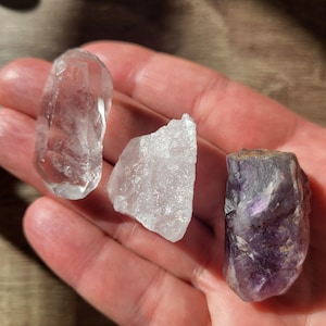 Puede incluir: Tres cristales en bruto: una punta de cuarzo transparente, un cúmulo de cuarzo transparente y un cúmulo de amatista púrpura.