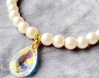 Può includere: Una collana di perle color crema con un pendente di cristallo iridescente a forma di goccia. Il pendente è avvolto in filo metallico color oro e pende da una fila di perle rotonde. La collana è esposta su una superficie grigia.