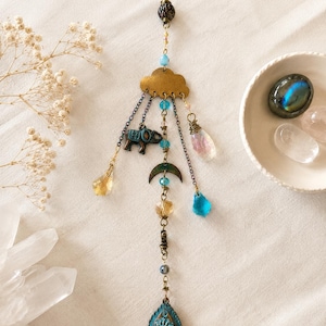 Op de afbeelding: Een hangend ornament met een koperen wolk aan de bovenkant, opgehangen aan een ketting. Het bevat bungelende elementen zoals een kleine olifantbedel, een halve maan, diverse blauwe en amberkleurige kralen en een druppelvormige hanger met bloemmotief.