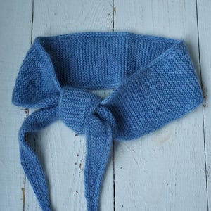May include: A blue knitted headband, tied in a knot. The headband is made of a soft, textured yarn and is a solid shade of blue. The headband is shown on a white wooden surface.