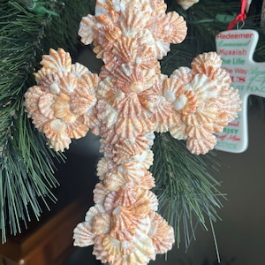 May include: A cross-shaped ornament made of seashells. The shells are arranged in a pattern to form the shape of a cross. The shells are a light brown color with white accents.