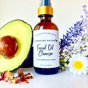 May include: A glass bottle with a gold lid and a white label that reads "HANDCRAFTED & SMALL BATCH EMBRACING NATURE Facial Oil Cleanser DRY/SENSITIVE SKIN Net 2 oz | 60 ml". The bottle is sitting on a white surface with an avocado half, dried rose petals, and a white flower with a yellow center.