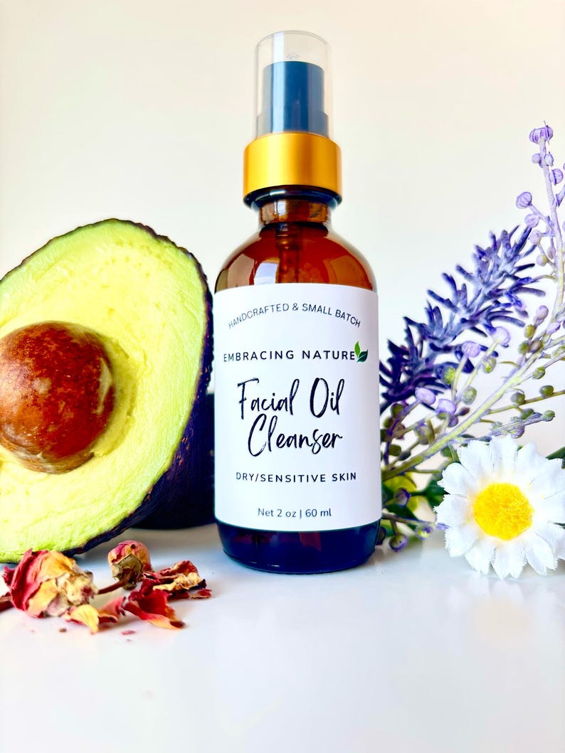 May include: A glass bottle with a gold lid and a white label that reads "HANDCRAFTED & SMALL BATCH EMBRACING NATURE Facial Oil Cleanser DRY/SENSITIVE SKIN Net 2 oz | 60 ml". The bottle is sitting on a white surface with an avocado half, dried rose petals, and a white flower with a yellow center.