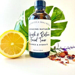 May include: A clear glass bottle with a black cap, labeled "Embracing Nature Refresh & Restore Facial Toner Balance & Hydrate Net 4 oz | 118 ml". The bottle is sitting on a white surface with a lemon slice and dried flowers.