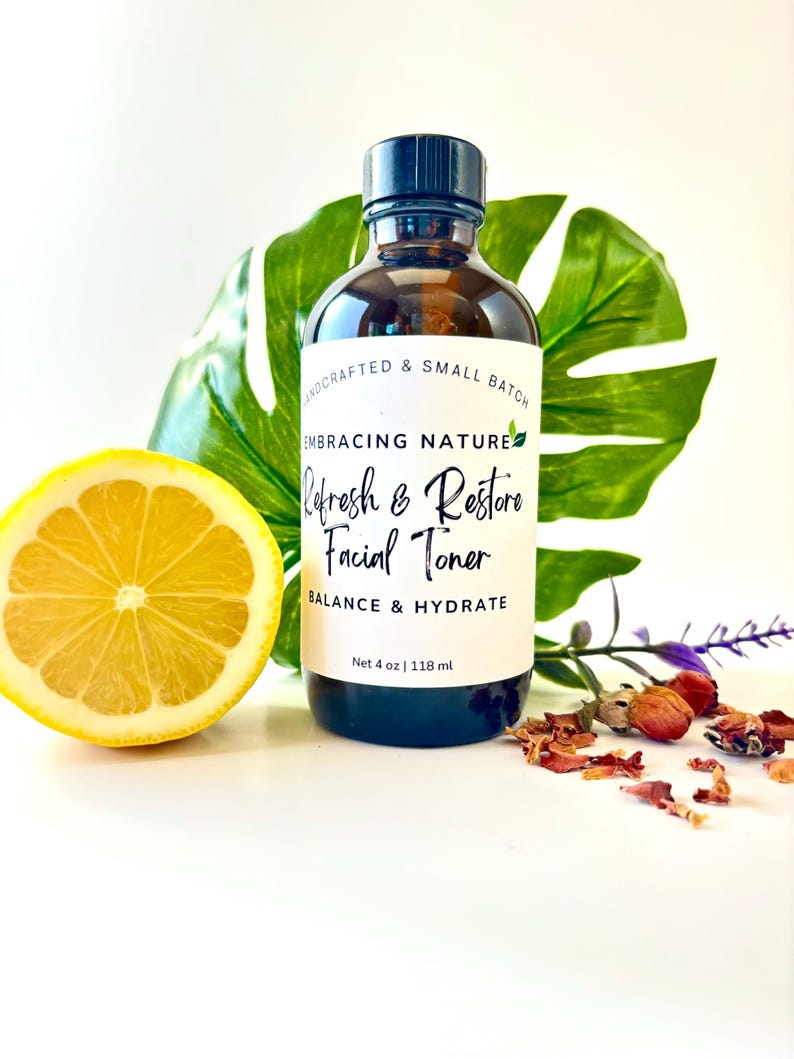 May include: A clear glass bottle with a black cap, labeled "Embracing Nature Refresh & Restore Facial Toner Balance & Hydrate Net 4 oz | 118 ml". The bottle is sitting on a white surface with a lemon slice and dried flowers.