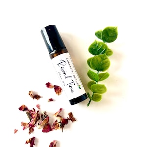 May include: A brown glass roll-on bottle with a black cap and a white label that says "Embracing Nature Rewind Time Roll On". The bottle is surrounded by dried rose petals and a sprig of green leaves.