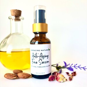 May include: A brown glass bottle with a gold lid and a clear dropper. The bottle is labeled "Anti-Aging Face Serum" and "Natural Timeless Glow". The bottle is sitting on a white surface with almonds, dried rose petals, and a lavender sprig.