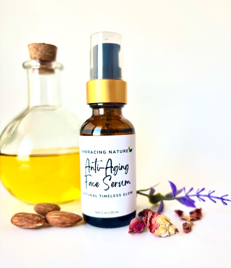 May include: A brown glass bottle with a gold lid and a clear dropper. The bottle is labeled "Anti-Aging Face Serum" and "Natural Timeless Glow". The bottle is sitting on a white surface with almonds, dried rose petals, and a lavender sprig.