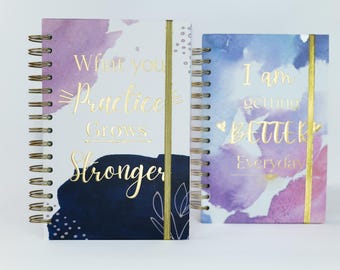 May include: Two spiral bound journals with watercolor designs and gold foil lettering. The first journal has a purple and blue design with the text "What you practice grows stronger". The second journal has a blue and pink design with the text "I am getting better everyday".