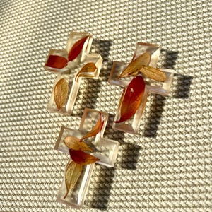 May include: Three clear resin crosses, each with dried leaves in red and brown. The crosses are on a textured, light-coloured surface, with shadows from the sunlight. The leaves are visible within the transparent resin.
