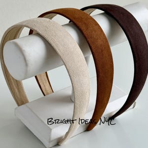 Può includere: Tre cerchietti con una consistenza morbida simile alla pelle scamosciata in color crema, marrone chiaro e marrone scuro. I cerchietti sono esposti su un supporto cilindrico bianco. Il testo BRIGHT IDEAS NYC è stampato in nero sulla base.