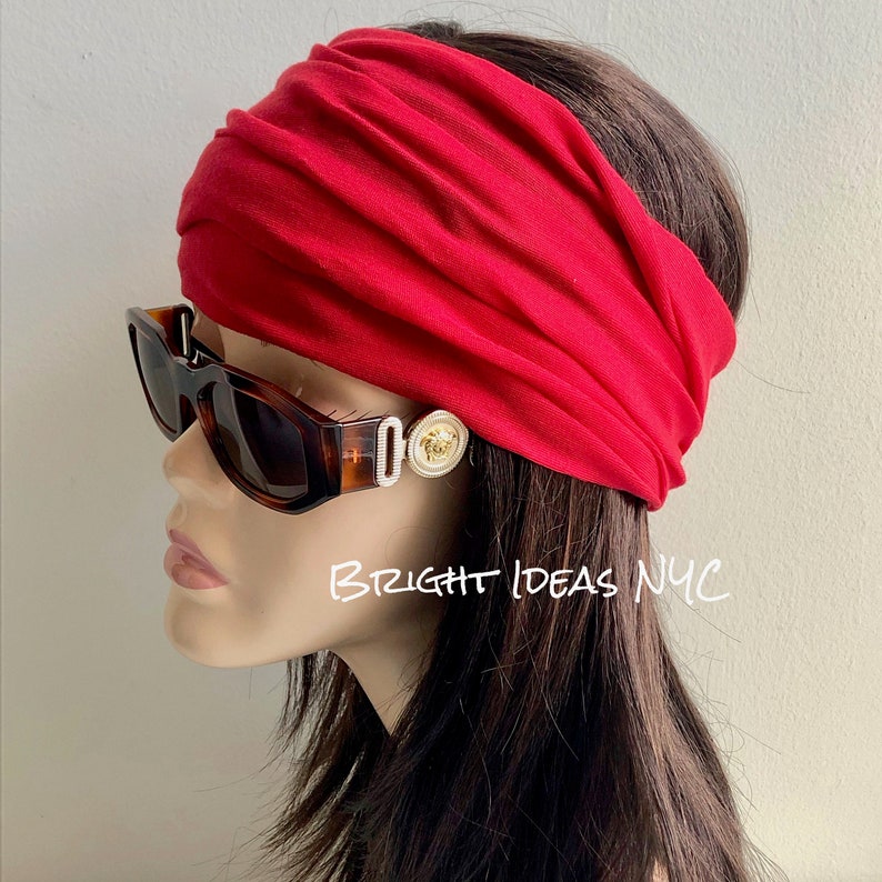 May include: A red fabric headband with a gold-toned decorative button. The headband is worn on a mannequin with brown hair and brown sunglasses.