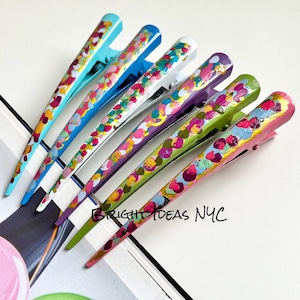 Könnte beinhalten: Satz mit sieben bunten Haarklammern mit einem gesprenkelten Farbmuster. Jede Klammer hat eine andere Farbe, darunter Blau, Grün, Lila, Rosa und Weiß. Die Klammern sind in einer Reihe angeordnet, wobei der Text "BRIGHT IDEAS NYC" in der unteren rechten Ecke sichtbar ist.