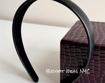 Puede incluir: Una diadema negra con un acabado liso y mate. La diadema está arqueada y se encuentra sobre una caja texturizada de color rojo oscuro. El texto "BRIGHT IDEAS NYC" es visible en la superficie.