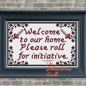 May include: A white cross-stitch design with a red and black border. The design reads "Welcome to our home. Please roll for initiative." and features two swords. The design is framed in a black frame. The text "The Debaucherous Needle" is below the design.