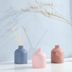 May include: Three ceramic diffuser bottles with wooden sticks. The bottles are in shades of blue, pink, and brown.