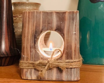 May include: A wooden candle holder with a cutout in the shape of a crescent moon. The holder is decorated with twine and has a small white candle inside. The candle holder is sitting on a wooden surface with a green vase and a brown vase in the background.