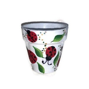 Puede incluir: Maceta de cerámica blanca con un diseño pintado a mano de mariquitas rojas, hojas verdes y pequeños puntos dorados. La maceta tiene una forma ligeramente acampanada y un borde gris oscuro. Decorativa, para interior o exterior.