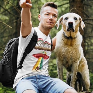 May include: A man wearing a white t-shirt with a retro-style graphic of a UFO and the word "Believe" in rainbow colors. He is sitting on a log in a forest with a dog beside him.