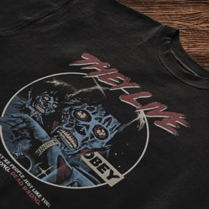 May include: A black t-shirt featuring a circular graphic with two blue-toned alien figures. The text "THEY LIVE" appears in red above the circle, and the word "OBEY" is printed inside. The text "YOU THINK THEY'RE PEOPLE JUST LIKE YOU. YOU'RE WRONG. DEAD WRONG." is printed in white and red.