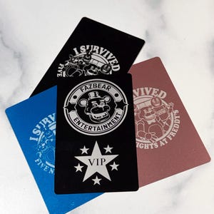 May include: Four rectangular cards with rounded corners. One black card features a white VIP star and the text "FAZBEAR ENTERTAINMENT". Another black card has the text "I SURVIVED FIVE NIGHTS". A blue card and a maroon card are also visible.