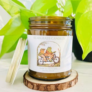 May include: A brown glass candle jar with a white label that says "Beargrass Candles" and features a cartoon illustration of two frogs riding a bicycle. The label also says "Made in Montana" and "9oz/Soy Blend".