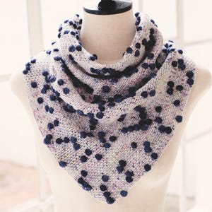 May include: A hand-knitted cowl scarf in a triangle shape, displayed on a mannequin. The scarf is a light gray with flecks of color and navy blue bobbles. The cowl is a cozy accessory.