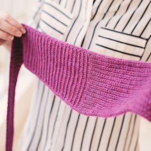 Könnte beinhalten: Ein gestrickter, magentafarbener Schal mit Rippenstruktur. Der Schal wird hochgehalten, um seine Länge und das detaillierte Strickmuster zu zeigen. Der Hintergrund ist ein gestreiftes Hemd.