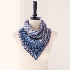 May include: A blue and tan crocheted scarf displayed on a mannequin. The scarf features a geometric pattern with a diamond-like design at the bottom. The color scheme includes shades of blue and tan, creating a textured appearance. The scarf is triangular in shape.