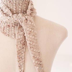 May include: A beige and brown crocheted scarf with a textured, knotted design. The scarf is draped over a mannequin, showcasing its triangular shape and long, pointed ends. The neutral color palette suggests a cozy, handmade accessory.