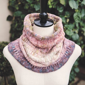 Puede incluir: Un cuello tejido a mano en tonos de rosa, morado y crema, exhibido en un maniquí. El cuello presenta un patrón de puntada texturizada y un borde acanalado. La paleta de colores incluye variaciones sutiles, creando una apariencia suave y mezclada.