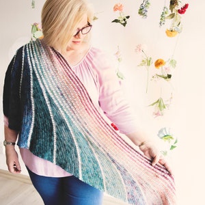 以下が含まれることがあります： ピンクのシャツと青いジーンズを着用した女性が、カラフルなクロシェのショールを持っています。ショールは、ティール、ブルー、ホワイト、ピンク、レッドなど、グラデーションカラーになっています。ショールは肩にかけられており、彼女は両手で持っています。
