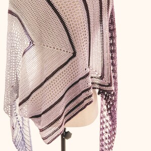 May include: A crocheted shawl with a purple and grey colour gradient. The shawl is made with a delicate openwork stitch and features stripes of a darker purple colour.