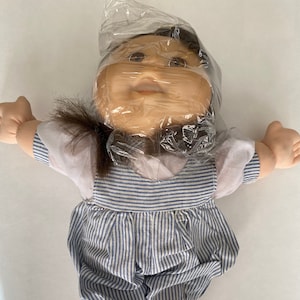 May include: A Cabbage Patch Kid doll with brown hair wearing a white shirt and blue and white striped overalls. The doll is wrapped in plastic.