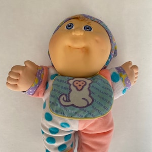 May include: A Cabbage Patch Kid doll wearing a pink and white polka dot outfit with a blue bib that says "Babyland Kid" in a repeating pattern. The doll has blonde hair, blue eyes, and a rosy complexion.