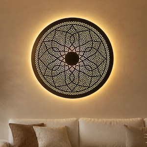 Op de afbeelding: Een ronde wandlamp met een zwart metalen frame en een ingewikkeld, lasergesneden geometrisch mandalapatroon. Het ontwerp bevat een massieve cirkel in het midden, omringd door herhalende bloemachtige vormen, verlicht door een warme gloed.
