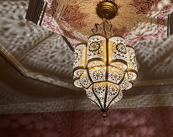 May include: Ornate, gold-colored Moroccan-style pendant lamp with intricate cut-out patterns, casting decorative shadows on a pink and white ceiling. The lamp is suspended from a decorative ceiling with carved details, creating a warm, inviting ambiance.