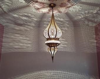 May include: A gold and white Moroccan pendant light fixture with intricate cutouts hangs from a ceiling with a star-shaped design. The light casts a pattern of shadows on the wall.